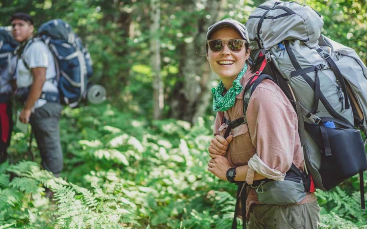 A person wearing a backpack smiles at the camera in a dense green forrest. There are other people wearing backpacks in the background. 